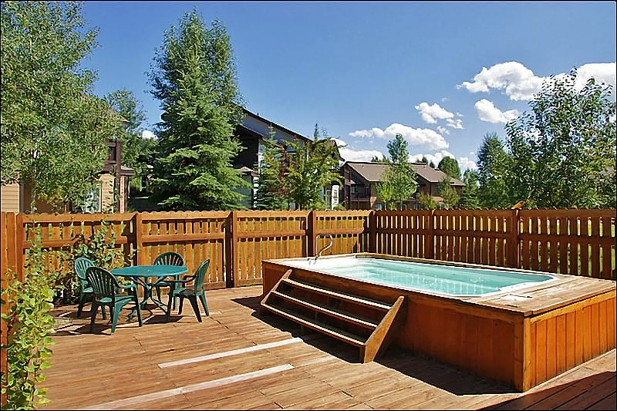 An outdoor wooden deck featuring a hot tub, table and chairs, and surrounding greenery. This tranquil setting is ideal for outdoor dining and social gatherings, with views that suggest a scenic backdrop.