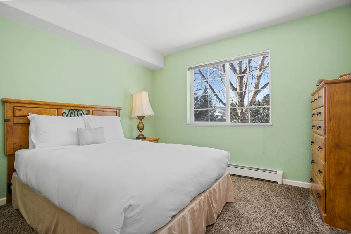 This image features a serene bedroom with a plush bed, a large window letting in natural light, a dresser, and a bedside lamp. The space projects a peaceful, restful atmosphere—ideal for a master suite retreat.