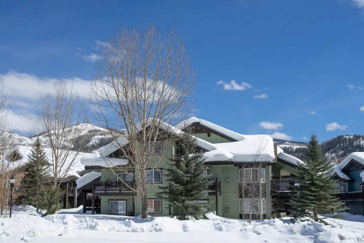 A snow-covered building surrounded by tall trees, with a tranquil winter scene that evokes the atmosphere of a mountain retreat. The green building reflects the cozy lodging set within a winter wonderland, while the visible balcony hints at relaxation after outdoor adventures.
