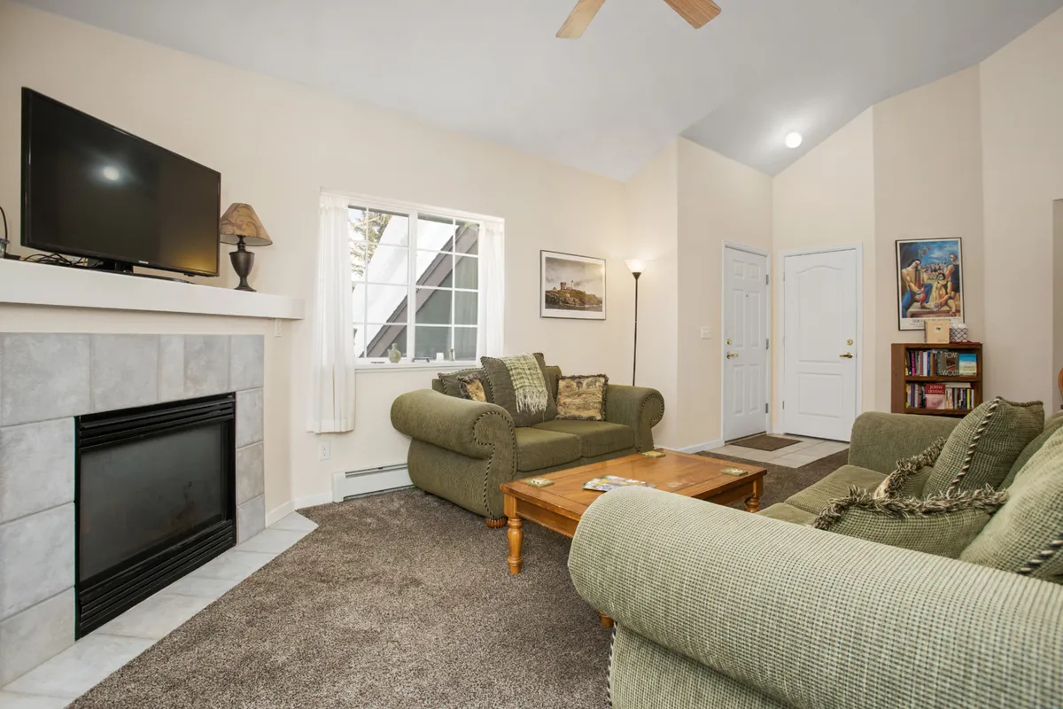 A comfortable living room with plush sofas arranged around a modern fireplace, a coffee table at the center, and a prominent wall-mounted HDTV. The room is filled with natural light, creating a cozy and welcoming environment ideal for relaxing, watching movies, or conversing with friends and family.