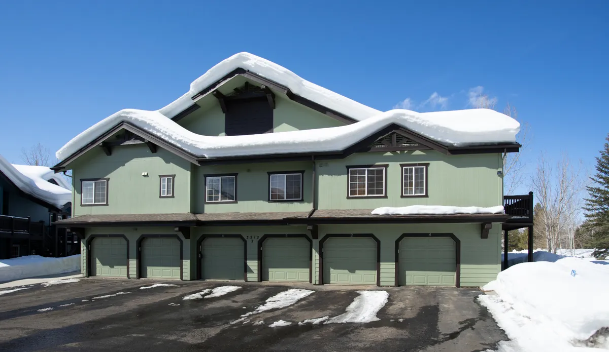 This image shows the exterior of the building with a snow-covered roof, green exterior siding, and prominent garage doors. The scene is set in winter, with snow blanketing the ground and trees, evoking a sense of preparedness for mountain living. The garage is clearly visible, highlighting the secure and practical storage for outdoor gear referenced in the section.