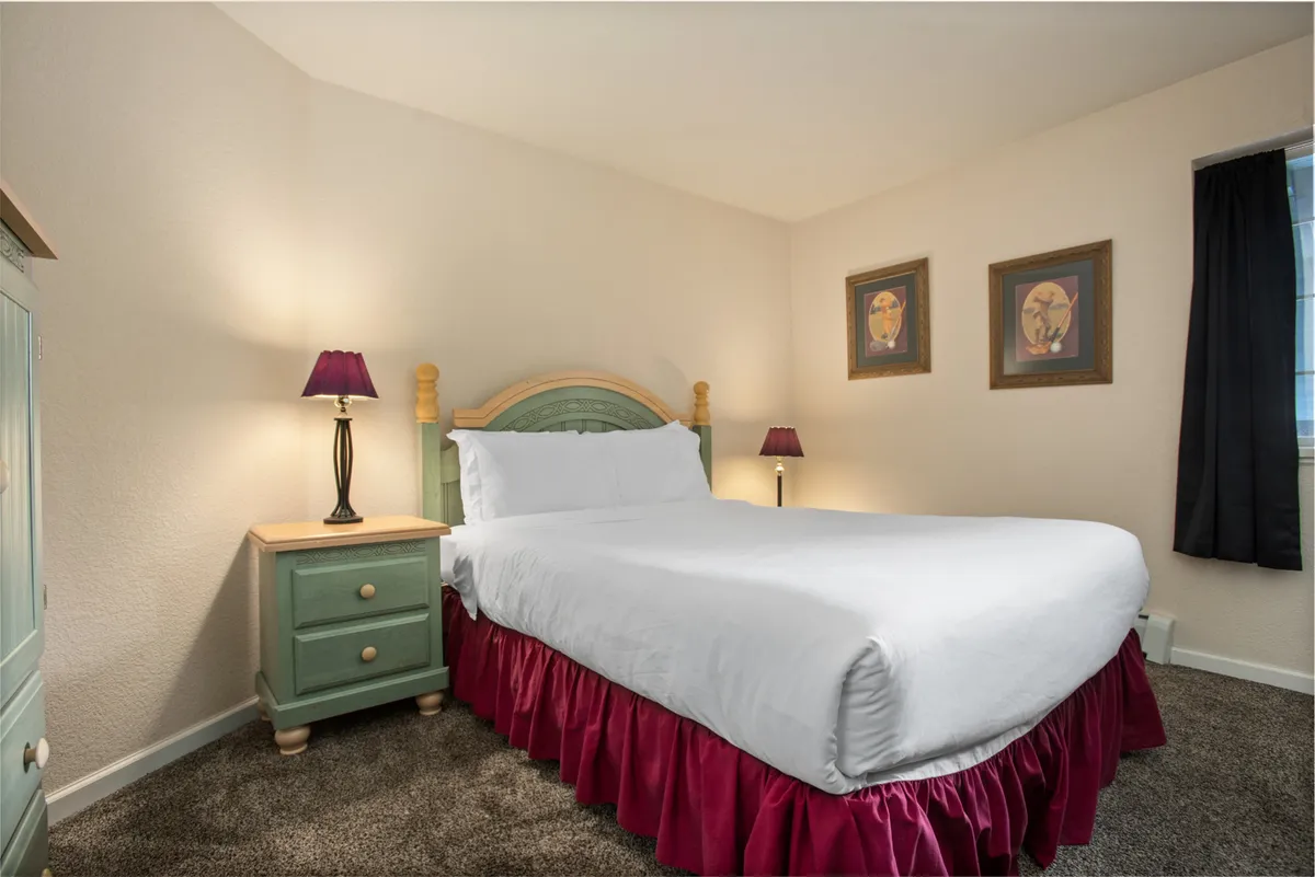 A cozy, inviting bedroom with a bed dressed in crisp white linens, a nightstand with lamps, and tasteful artwork. The soft lighting and tidy arrangement create a tranquil setting.
