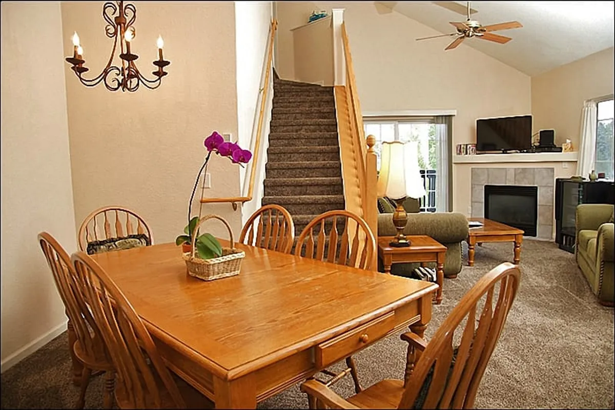 A spacious open-concept living and dining area featuring a large dining table, a cozy sofa, a fireplace, a staircase, and a decorative chandelier. The space is bathed in warm light and feels perfect for gatherings, with direct visual connection between the living and dining zones.