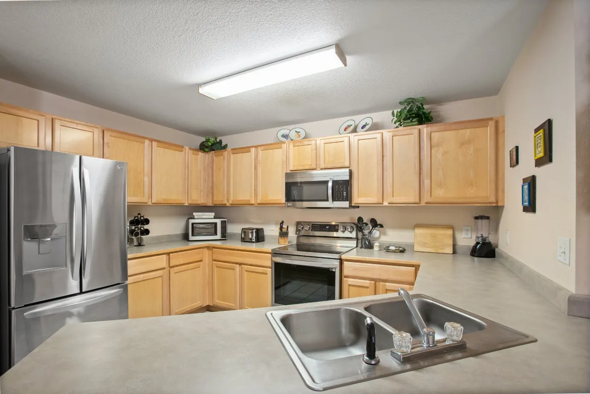 A warmly lit kitchen featuring a stainless steel refrigerator, microwave, sink with faucet, and wooden cabinets. The spacious counters suggest ample room for cooking and meal preparation, supporting both everyday living and entertaining. The environment feels inviting and functional for longer stays, with easy access to full kitchen amenities.
