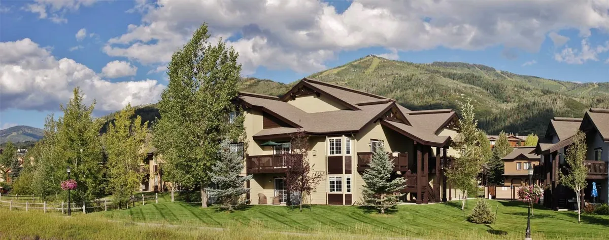 The hero image showcases the exterior of a modern condo nestled among lush trees, with dramatic mountain peaks in the background. The building’s architecture blends harmoniously with the natural landscape, while the balcony hints at spectacular sunset views. The serene setting invites moments spent enjoying wildlife sightings and outdoor gatherings, perfectly embodying the spirit of Steamboat Springs.